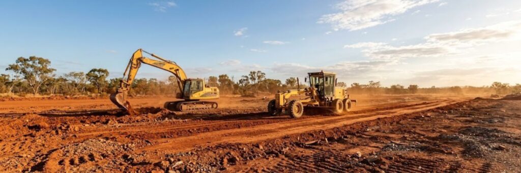 North Queensland civil earthworks site — excavator and motor grader on red laterite earth at golden hour